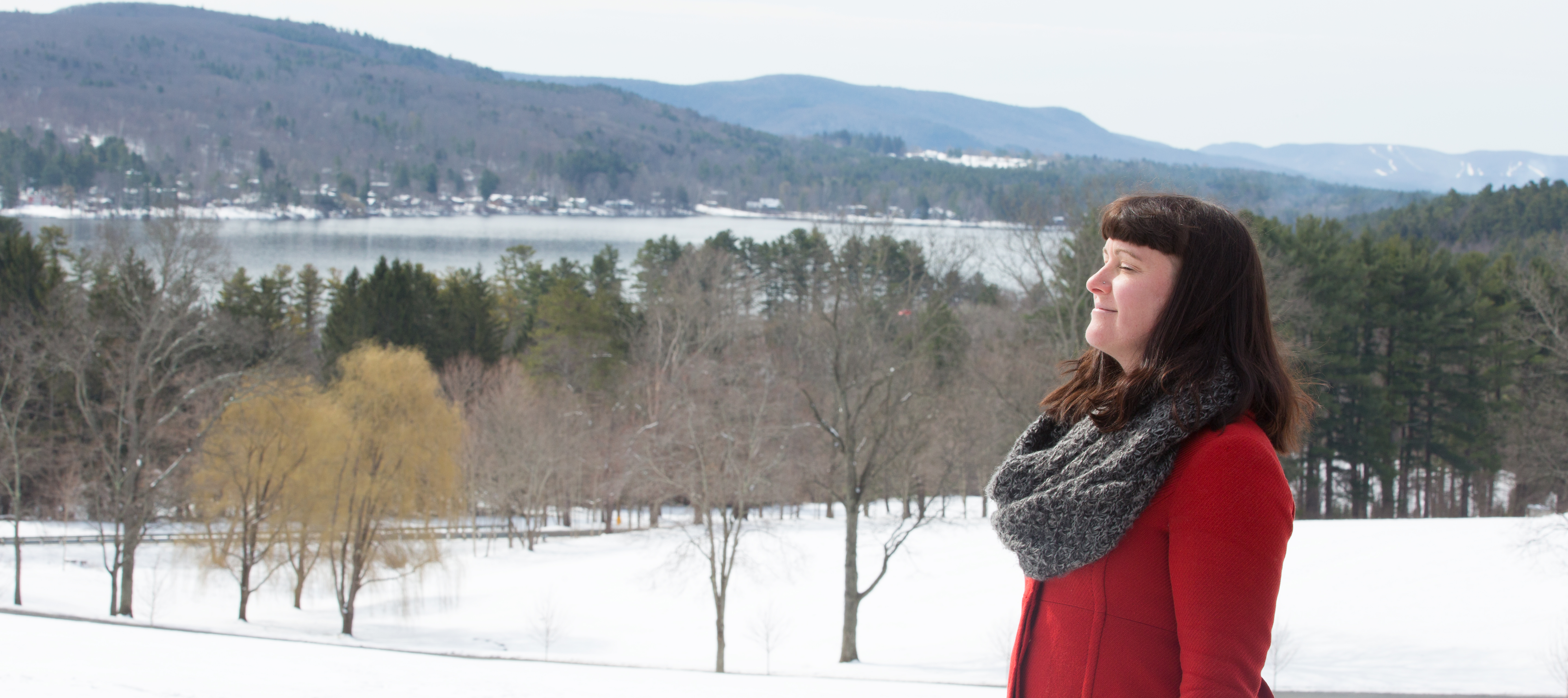 Woman standing with eyes closed outdoors in the winter with hills and lake in the background.