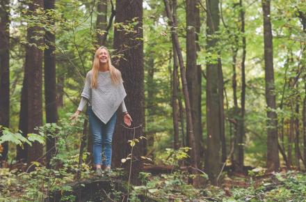 Smiling woman with arms lowered and eyes closed standing in the woods.