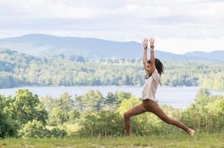 Woman with hands raised in a yoga pose next to a lake and mountains in the Summer