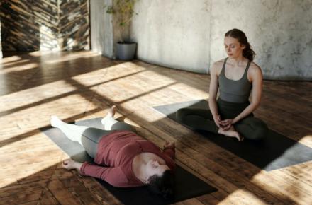 One woman seated on a yoga may, one woman lying on a yoga mat.