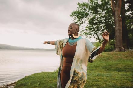 Black woman standing at the edge of a lake with eyes closed and arms outstretched.