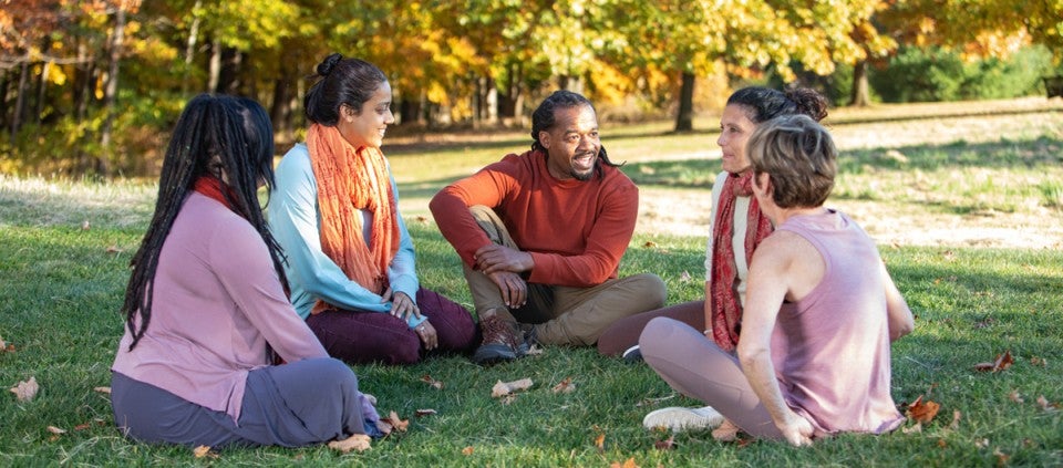 A diverse group of people stand in a field in Fall, their hands outstretched to the sky.
