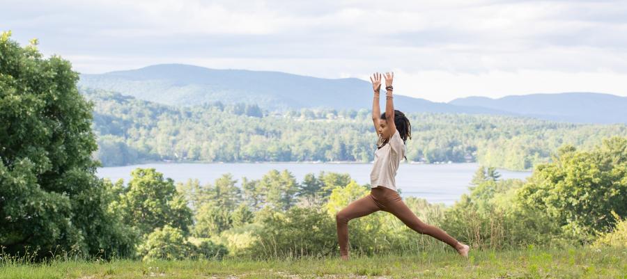 Woman with hands raised in a yoga pose next to a lake and mountains in the Summer