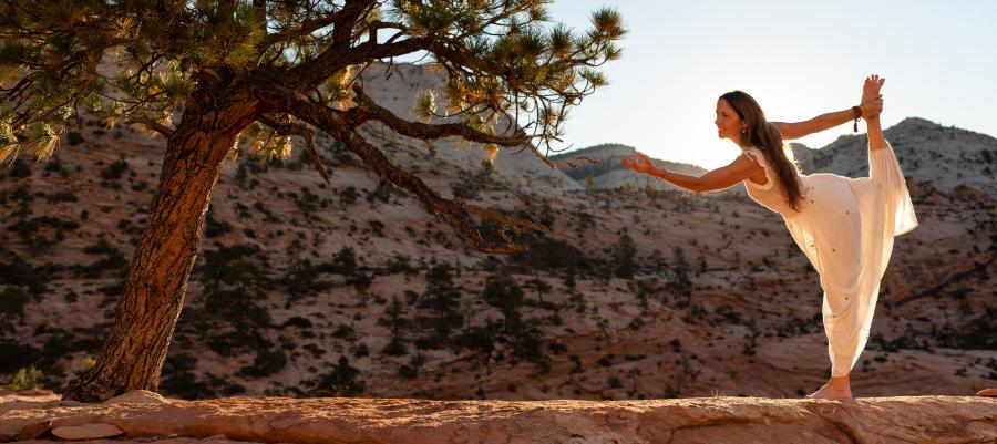 Kia Miller practicing yoga outdoors in a desert landscape next to a tree, with hills in the background