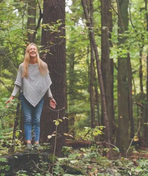 Smiling woman with arms lowered and eyes closed standing in the woods.