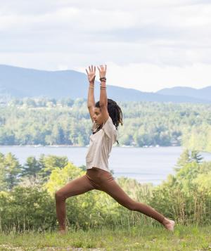 Woman with hands raised in a yoga pose next to a lake and mountains in the Summer