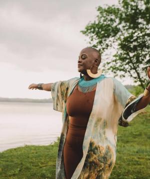 Black woman standing at the edge of a lake with eyes closed and arms outstretched.
